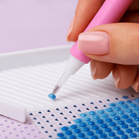 Close-up van een diamond painting pen die een blauw steentje plaatst op een canvas met symbooltjes.