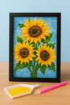 Diamond painting van drie zonnebloemen in zwarte lijst met applicatiepen en bakje gele steentjes op houten tafel
