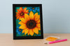 Diamond painting van zonnebloemen in een zwarte lijst met applicatiepen en bakje steentjes op een houten tafel